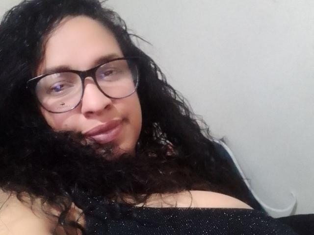 ZoehFerraro, com cabelos pretos cacheados, óculos e um leve sorriso, exibindo seus seios grandes, vestindo uma blusa preta brilhante, em um fundo simples..