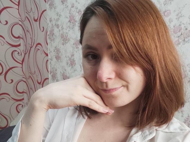 LeonoraGrego with reddish-brown hair, fair skin, and a subtle smile, resting her chin on her hand, wearing a white shirt, showing her small, cute nose and soft tits.