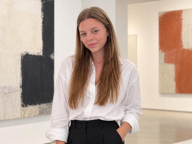 RhonaBells with long brown hair, white button-up shirt, black high-waisted pants, hands in pockets, slight smile, standing in a modern art gallery, looking relaxed.