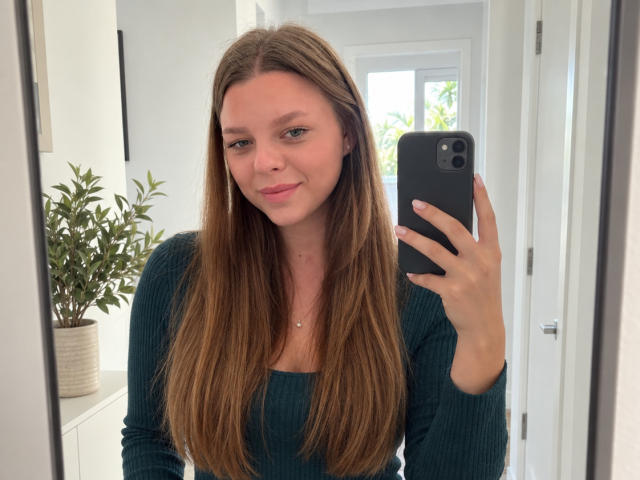 RhonaBells with long brown hair, green eyes, and a subtle smile, wearing a teal rib-knit top, taking a mirror selfie with a black phone, showing a small nose piercing.