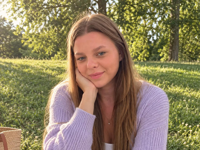 RhonaBells with long brown hair, purple sweater, and pink lips, resting her head on her hand, smiling softly in a sunlit grassy area. She has small tits.
