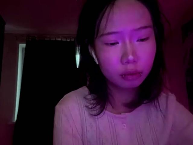 SanjuanitaCejka with purple-lit face, dark hair, closed eyes, and slightly open mouth, wearing a white shirt, looking down, in a dimly lit room.