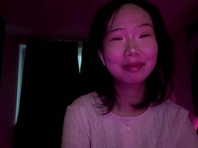 SanjuanitaCejka with dark, straight hair, pinkish-purple light on face, smiling slightly, wearing a white, button-up shirt with subtle patterns, in a dimly lit room.