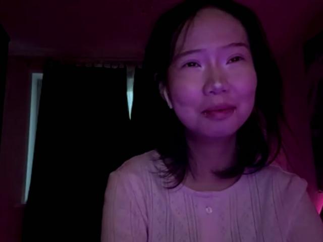 SanjuanitaCejka with a slight smile, purple-tinted skin, black shoulder-length hair, wearing a white button-up shirt, dimly lit, upper body, looking slightly to the left.