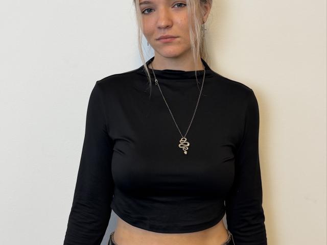 AshlieKeziah mit blonden Haaren, einem schwarzen, langärmligen Crop-Top, das ihre kleinen Brüste zeigt, trägt eine Schlange als Halskette und steht vor einer schlichten Wand..