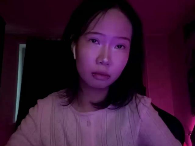 SanjuanitaCejka has medium-length black hair, wearing a white, ribbed, long-sleeve shirt, with purple lighting, looking slightly open-mouthed. She has earbuds in.