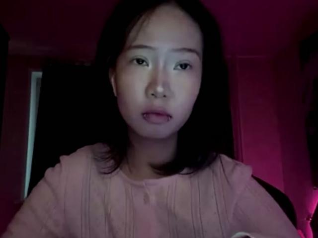 SanjuanitaCejka with straight black hair, pink ruffled shirt, and a slightly open mouth showing a small tooth, in dim pink-lit room. She has a nervous or surprised expression.