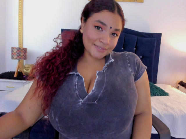 DarsshaHara with curly red hair, wearing a gray, velvet, v-neck shirt, showing her large, prominent tits. She's smiling, standing in a bedroom, with a dark blue headboard.