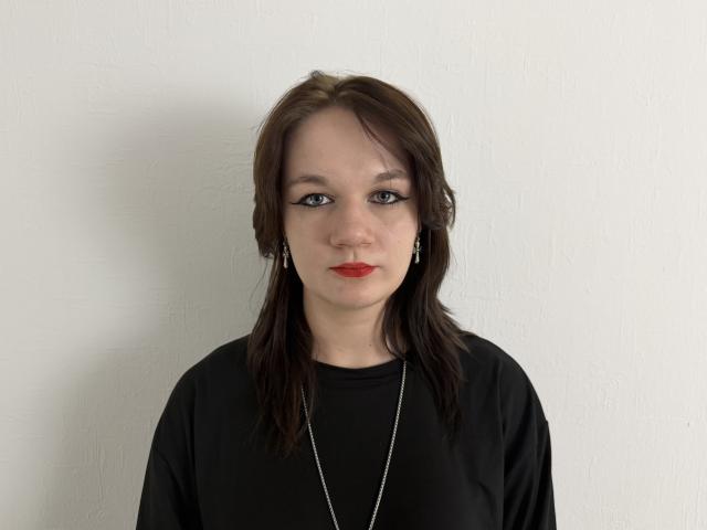 Eliana Laneve hat glattes, braunes Haar, roten Lippenstift, schwarzen Eyeliner, ein schwarzes Hemd und eine silberne Halskette. Sie schaut mit einem neutralen Gesichtsausdruck in die Kamera..