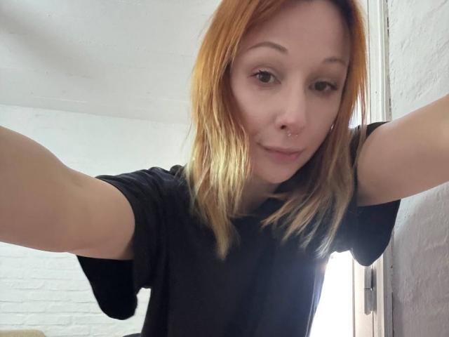 HannaCool mit hellbraunen Haaren, schwarzem T-Shirt, Nasenring und einem leichten Lächeln, macht ein Selfie mit ausgestreckten Armen und zeigt dabei ihre Brüste leicht..