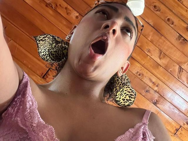 NatalyBlonder with her mouth open, wearing leopard print earrings and a pink lace top, taken from a low angle. Her pink lace top reveals her small, perky tits.