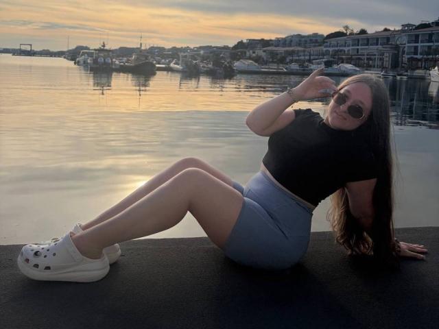 NikkaVeira sits on a dock, legs spread, wearing a black top, blue shorts, white sneakers, and sunglasses, drinking from a cup, with a calm waterfront at sunset.