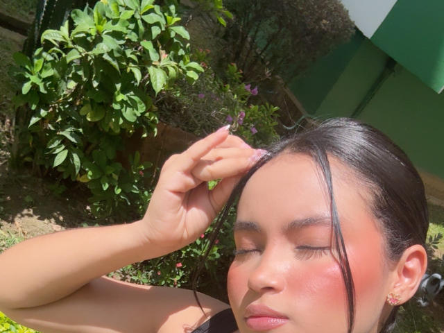 MiiaPalmer with her eyes closed, holding a tiny purple flower against her forehead, her pinkish-red cheeks glowing in sunlight, black hair pulled back, wearing a black top.