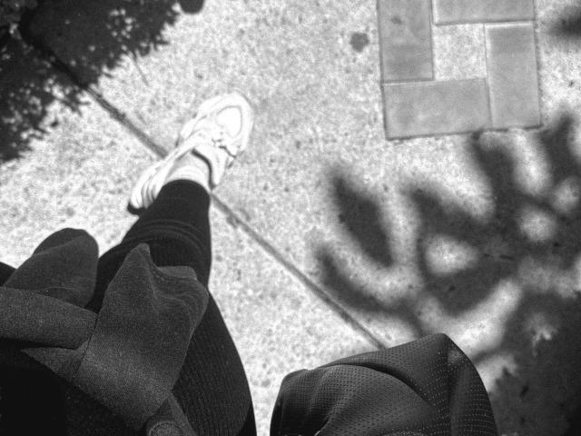 MiiaPalmer's foot in a white sneaker with laces, black pants, and a black backpack, shadowed on a concrete sidewalk with a brick pattern in the top right.