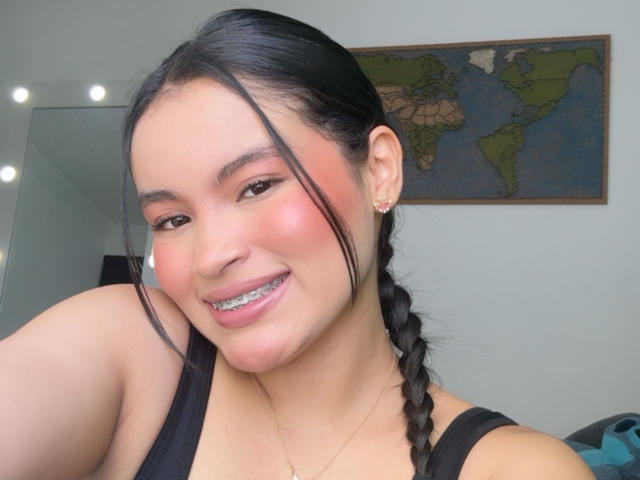 MiiaPalmer with black braids, pink blush, silver braces, smiling, black tank top, and earrings, showing off her cute face and light brown skin. World map in background.