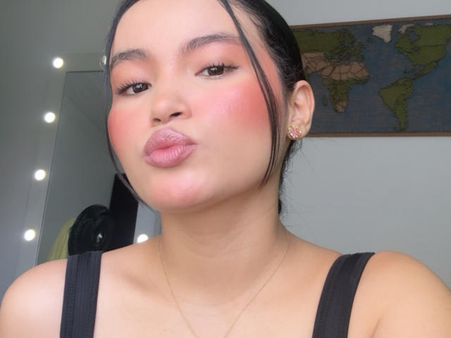 MiiaPalmer with pink blush, pouty lips, black hair in a low bun, wearing a black top, small gold earrings, and a thin necklace, in a selfie.