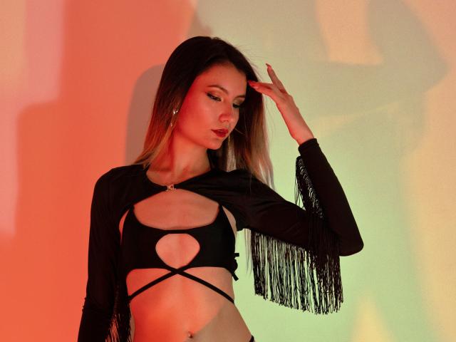 AlanisBlanc with long blonde hair, wearing a black fringed crop top with cutouts, exposing her small tits and toned belly, touching her head, in a red and green light.