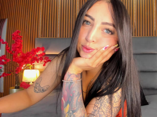 AnastasiaVolkov has long black hair, blue eyes, and is resting her face on her hand with pink nails. She has tattoos on her arm and a bracelet. Boobs visible.