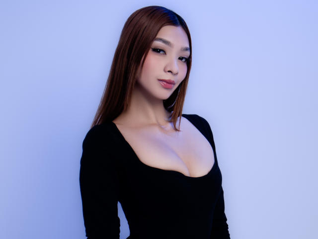SofiGale, com cabelo castanho, blusa preta de manga comprida e seios grandes visíveis, está em pé em frente a uma parede azul, olhando ligeiramente para o lado com um sorriso discreto..