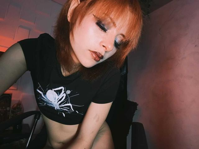 ViolettClay met oranje haar, zwarte oogmake-up en een spinnen-shirt, leunt voorover, waarbij haar kleine borsten en platte buik zichtbaar zijn, met een verleidelijke, lichtjes fronsende uitdrukking..