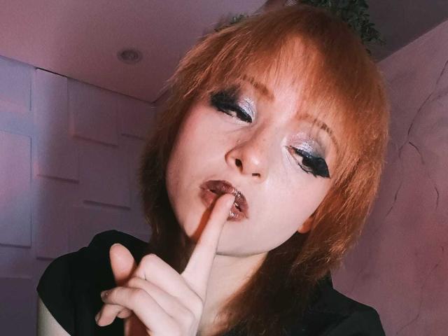 ViolettClay met rood haar, bleke huid en blauwe glitterschaduw, die op haar vinger zuigt, met zwarte eyeliner en licht open lippen. Ze draagt een zwarte top..