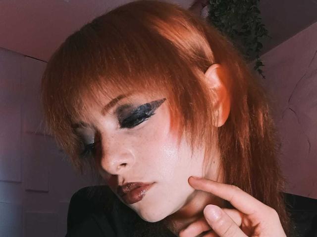 ViolettClay met rood haar, donkerblauwe winged eyeliner, glanzende roze huid, sproeten en glanzende bruine lippenstift, die haar kin aanraakt met haar vingers..