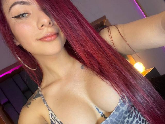 ValeryHarllow with long red hair, subtle eyeliner, and glossy pink lips, wearing a leopard-print top, showing her tits and tattoos, holding up her arm.