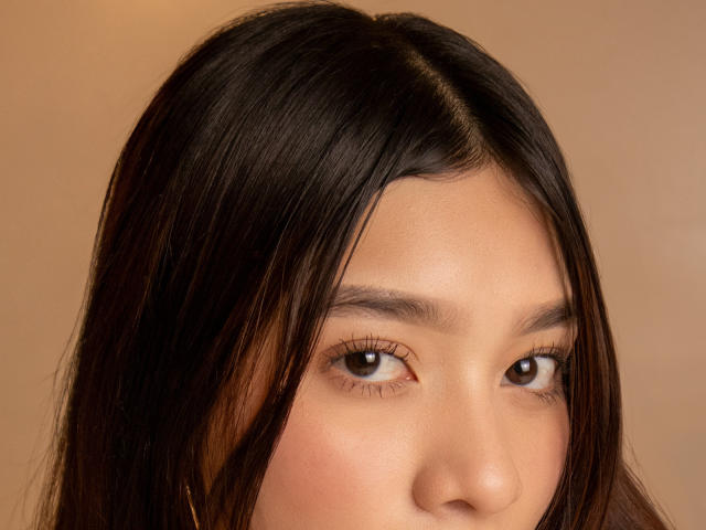 Het gezicht van SofiGale, bruine ogen, lang zwart haar, licht open lippen, gladde huid, subtiele make-up, warme bruine achtergrond, zacht licht, zachte uitdrukking, kleine gouden oorbellen aan de linkerkant..