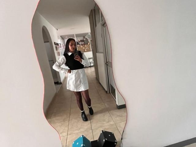 PaulaLoughty stands in a mirror, wearing a black top, white skirt, maroon pants, and black boots, with a hand on her hip, looking at her phone.