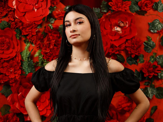 DevineSimone, con lunghi capelli neri, indossa una maglia nera con una spalla scoperta, orecchini a catena dorati e una collana, e si presenta con sicurezza, mani sui fianchi, con delle rose rosse sullo sfondo..