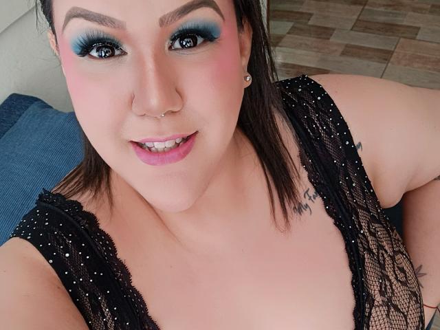 GiiannaMorrys with blue eye makeup, pink blush, and pink lipstick, wearing black lace top, showing cleavage, nose piercing, and smiling. Her tattoo is partially visible on her right shoulder.