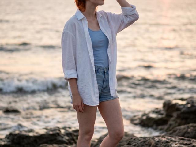 IngaLeerar, con cabello rojo, vestida con una camisa blanca de botones sobre un top azul y pantalones cortos vaqueros, de pie junto a la costa rocosa del océano, con la mano cerca de su cabeza..
