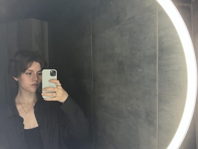 HanhFontillas met kort bruin haar, die een witte telefoon vasthoudt en een spiegel-selfie maakt, draagt een zwarte blouse, een ketting en een ring, in een donkere betegelde badkamer..