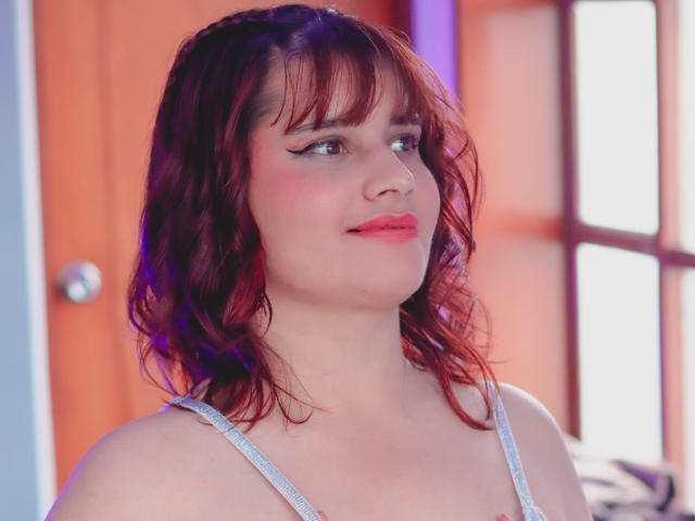VivianFranco has red curly hair, white eyeliner, pink lipstick, and is looking to the side with a slight smile. She's wearing a white strap top, showing her tits.