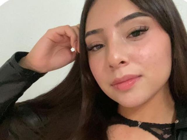 GigiOchoa a de longs cheveux bruns, des sourcils épais et des lèvres roses brillantes. Elle porte un haut en cuir noir et touche son oreille avec un boucle d'oreille en argent. Ses seins sont partiellement visibles..
