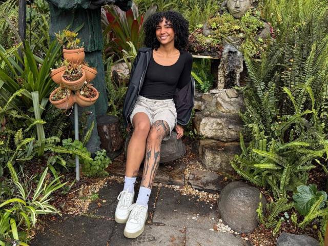 CinthiaWalker with curly black hair, black top, beige shorts, white socks, and white sneakers, sitting, showing tattoo on right thigh, smiling, surrounded by green plants and rocks.