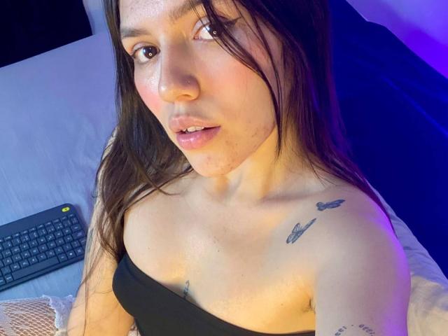 SophiaMarx with long brown hair, pink lips, and small tits, wearing a black top, showing two bird tattoos on her shoulder, sitting with a keyboard in the background.