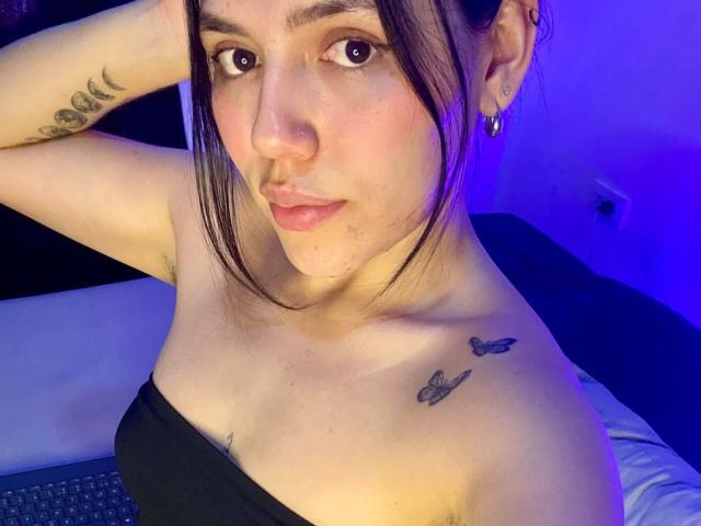 SophiaMarx with brown eyes, black strapless top, tattoos on shoulders, ear piercings, and purple blue lighting, looking at camera with hand in hair. Boobs exposed, slight pink blush.