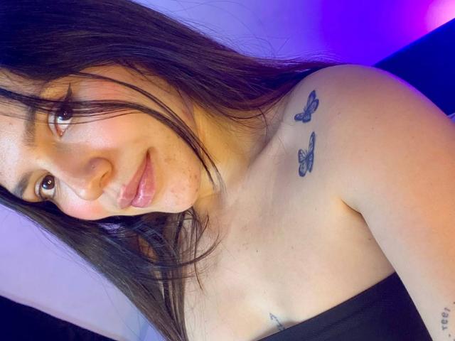 SophiaMarx with long brown hair, light skin, small tits, butterfly tattoos on shoulder, slight smile, pink lips, subtle makeup, and a black strapless top, in a selfie.