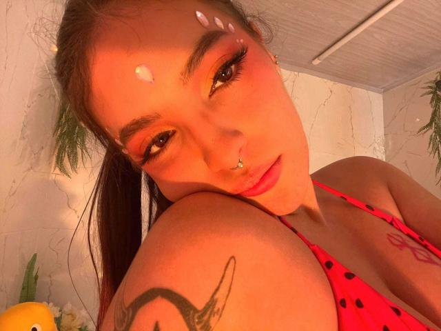 KatarinaNovak with cum on her forehead, red polka dot bikini, tattoos on her shoulder, and nose ring, looking at the camera with orange eye makeup.