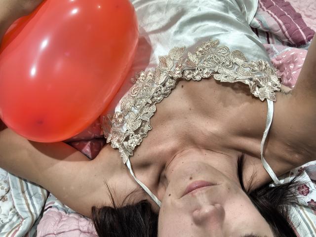 OrianaLaFrancaise liggend op bed met een rode ballon, een witte kanten top, armen omhoog, ogen gesloten, donker haar, bleke huid, kleine sproeten op de nek, zacht licht, ontspannen uitdrukking..