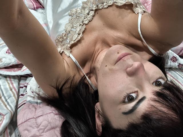 OrianaLaFrancaise, deitada em uma cama, vestindo um vestido branco de renda, olhando para cima, cabelos escuros, olhos castanhos, pequenas sardas no rosto, alças brancas, colcha rosa e branca..