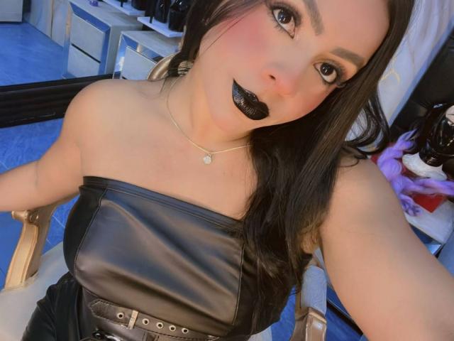 LindaFrancoo trägt ein Kleid mit schwarzem Ledergürtel, hat schwarzen Glitzer-Lippenstift, langes dunkles Haar, sitzt auf einem Stuhl, hat blaue Augen, helle Haut, eine Halskette und einen schwarzen Gürtel. Im Hintergrund ist ein Spiegel zu sehen. Der Stuhl ist aus Holz..