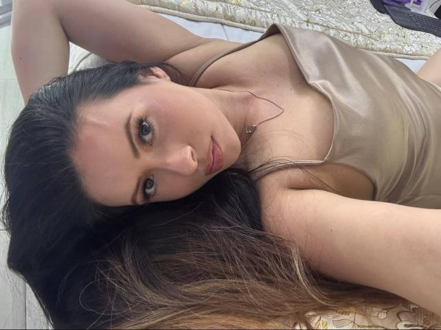 HannaRobertss lying on a bed, wearing a beige tank top, arms above head, brown hair, green eyes, looking at camera, subtle makeup, necklace, soft light, relaxed.