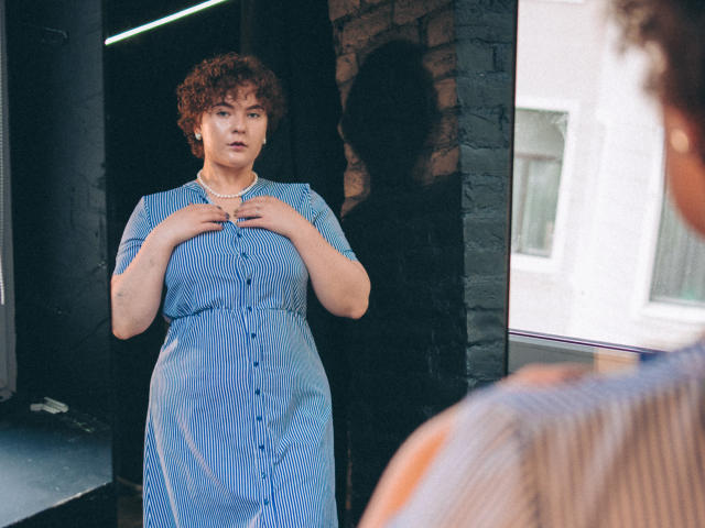 MelittaAdams stands in a blue, buttoned dress with short curly hair, holding her chest, shadowed on a black brick wall, looking at a blurred figure in the foreground.