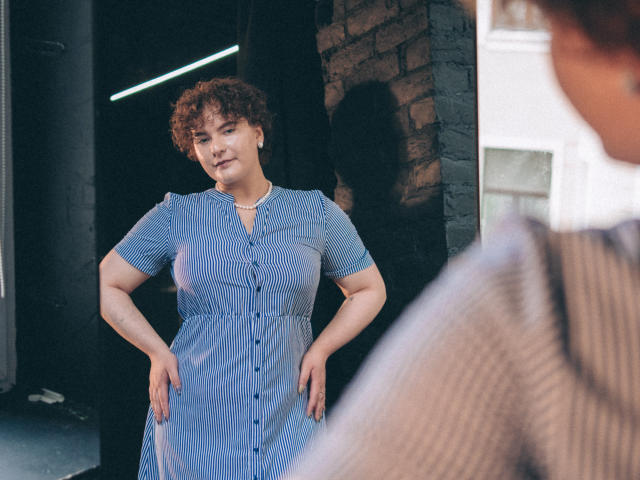 MelittaAdams, curly redhead, wearing blue striped dress, hands on hips, standing, reflection in mirror, blurry man in foreground, soft light, casual urban setting, confident expression.