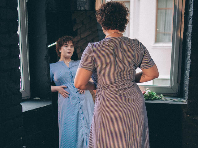 MelittaAdams in a blue dress stands facing a mirror, her back to the camera, showing her ass in a striped dress, curly brown hair, hands on her belly.