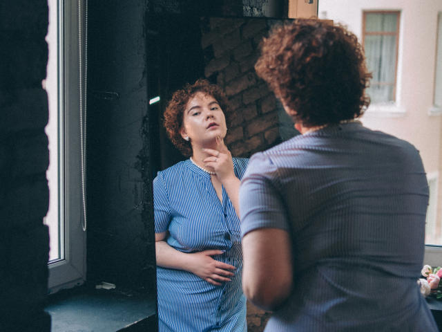 MelittaAdams with curly brown hair, blue striped shirt, and hand on belly, stands in front of a mirror, touching her chin, reflecting her serious, thoughtful expression.