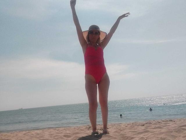 Knoha se tient sur la plage, les bras levés, vêtue d'un maillot de bain une pièce rouge, d'un chapeau de soleil et de lunettes de soleil, exposant ses fesses et ses petites seins sous la lumière vive du soleil..