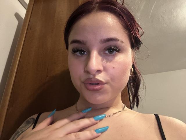 Luucie with puffy lips, blue nails, tattoo on shoulder, wearing black strap, red hair, green eyes, gold hoop earrings, and a chain necklace, looking at camera.